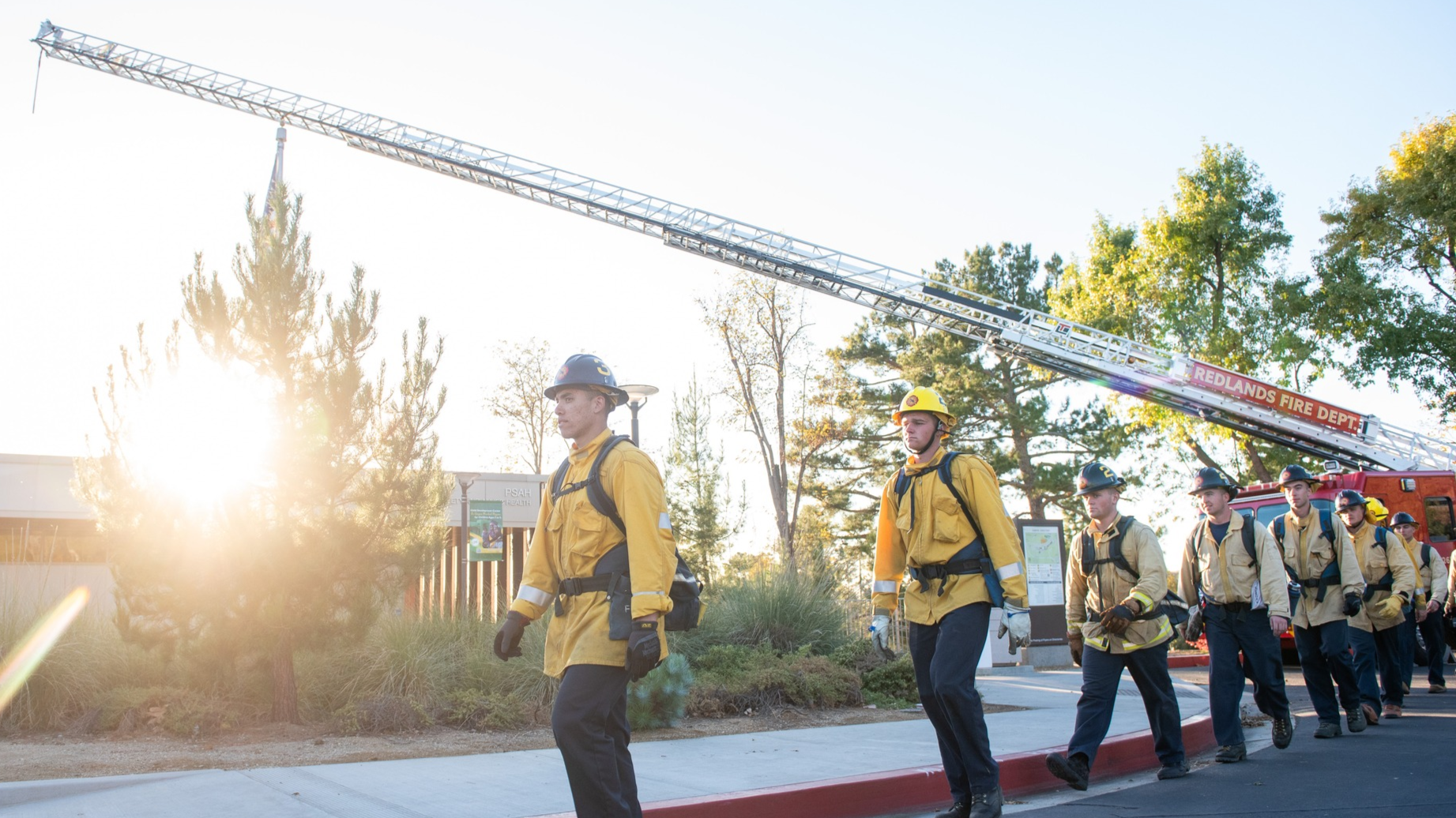 Fire academy cadets in yellow turnout gear marching beneath an extended fire department ladder truck.