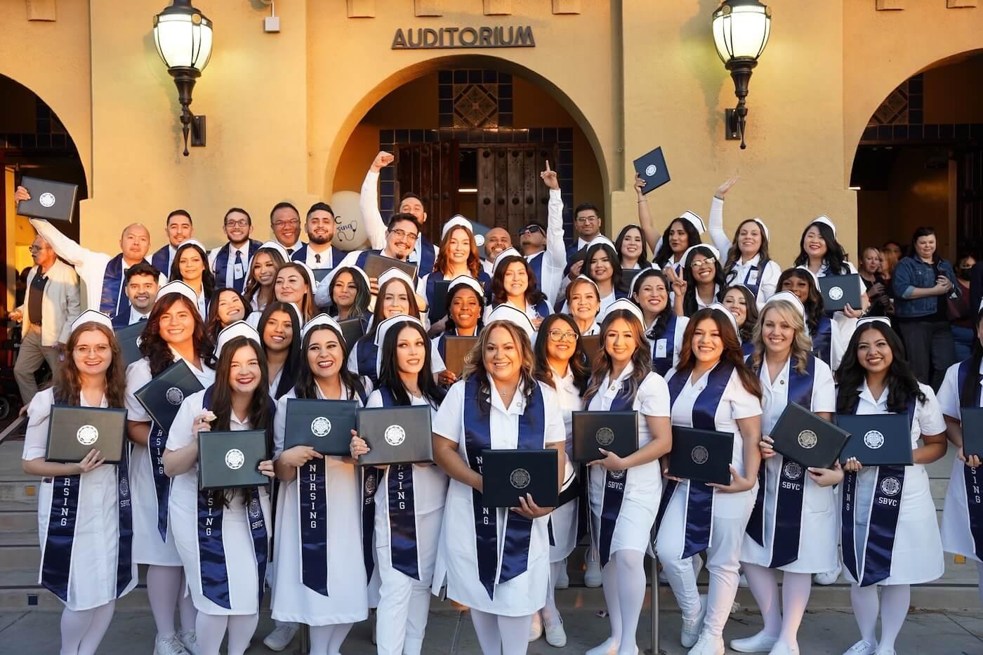San Bernardino Valley College nursing graduates in white uniforms and SBVC nursing stoles celebrating on the steps of the campus auditorium.