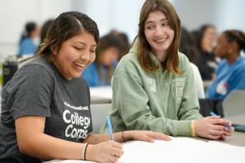 Two smiling students holding pens sitting next to each other in classroom setting.