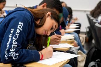 Students seated in a classroom, writing on papers with clipboards attached to chairs.